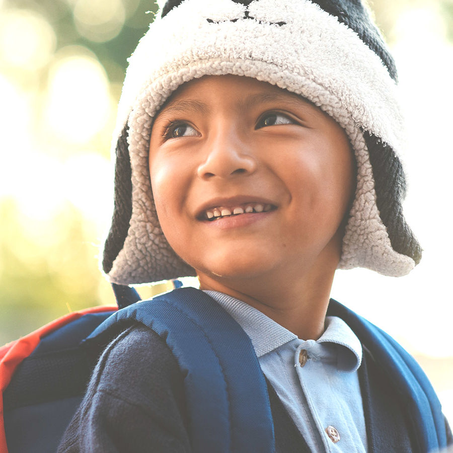 Kid with a fuzzy, warm hat and school uniform complete with backpack smiles